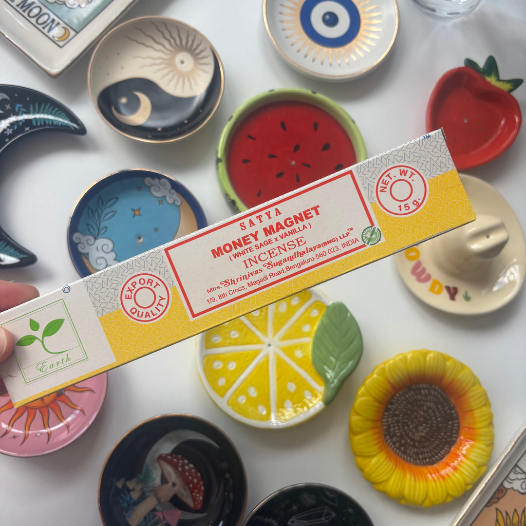 Box of Satya Money Magnet Incense Sticks with decorative plates in the background