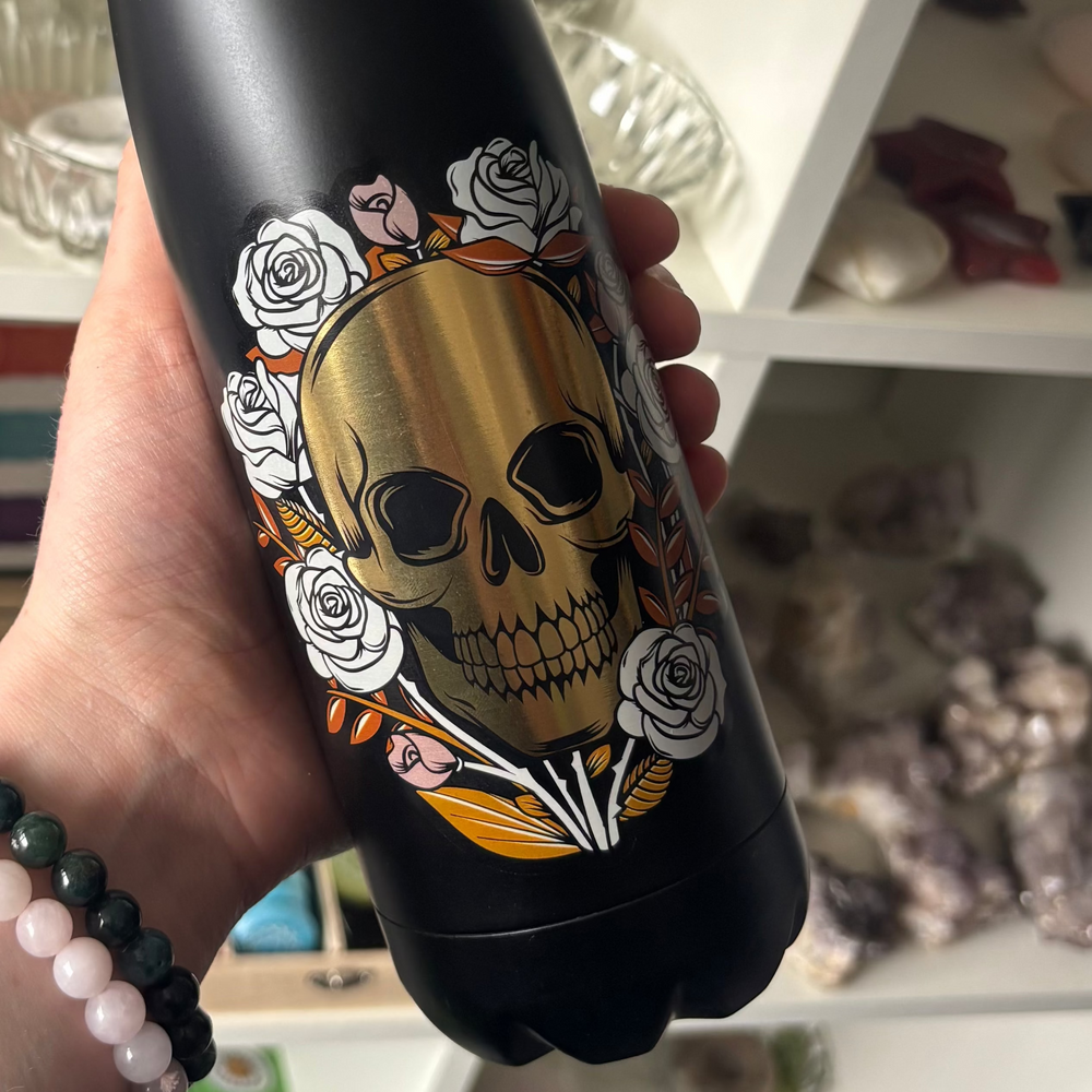 Black water bottle with skull and floral design held by a person in front of shelves.