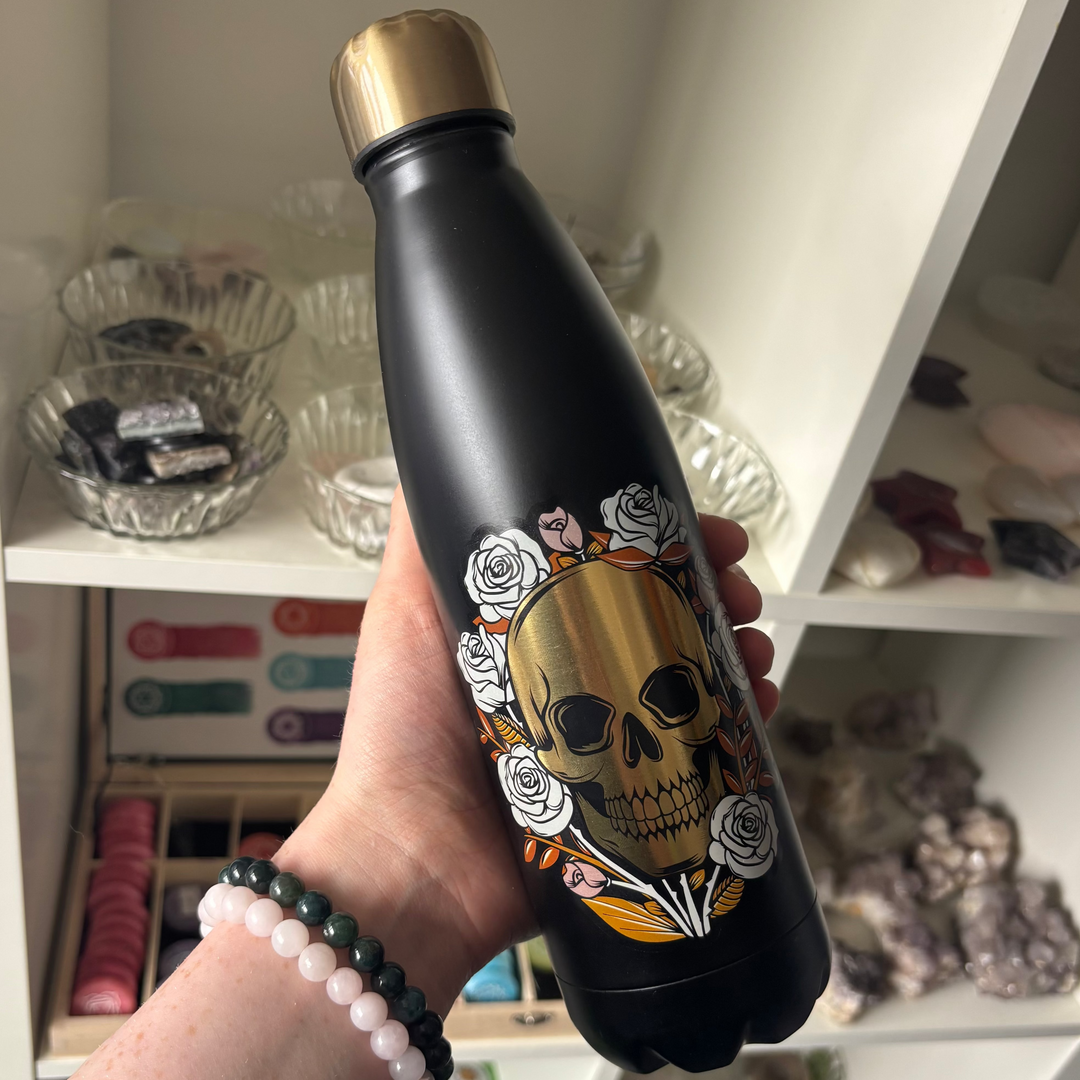 A hand holds a matte black insulated bottle featuring a large gold skull design surrounded by white roses and orange accents, with a blurred shelf of crystals in the background.