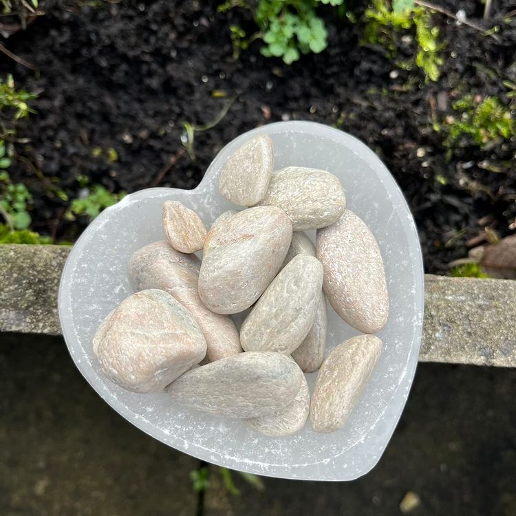 A heart-shaped translucent white stone bowl filled with smooth, polished river stones in light beige and gray tones, photographed outdoors against a dark soil background.