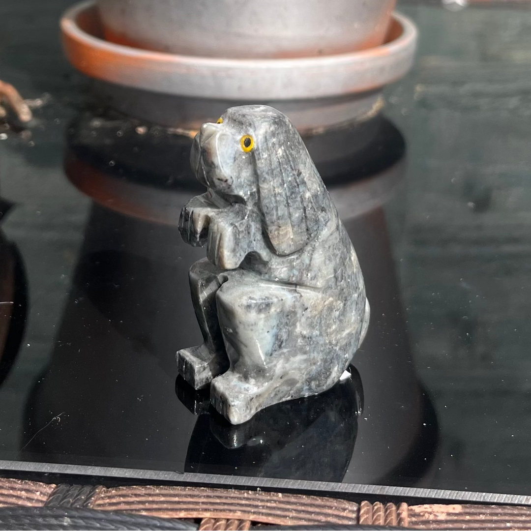 A soapstone dog carving sculpture, placed on a windowsill with a bird figurine in the background, next to a potted plant.