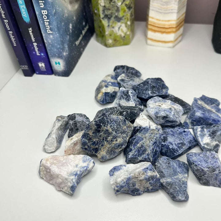 A pile of rough blue and white Raw Sodalite Crystal Pieces on a white surface, with a few books visible in the blurred background.