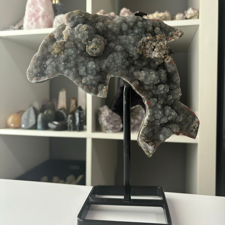 A Sphalerite Dolphin Carving on a black stand, displayed in a room with various crystals and rocks.