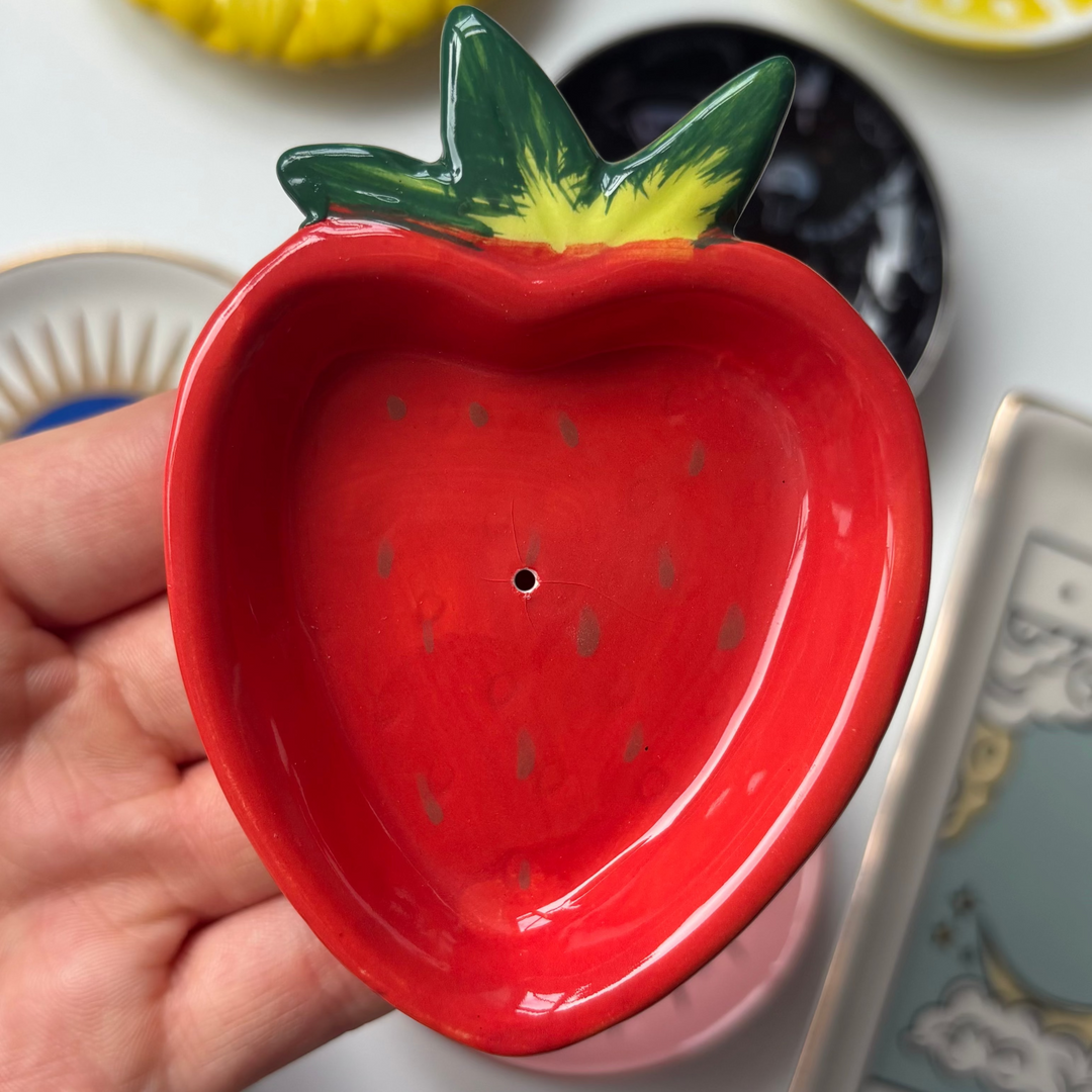 Red ceramic strawberry-shaped Strawberry Incense Stick Holder held by a hand with various small ceramic items and a tarot card in the background.