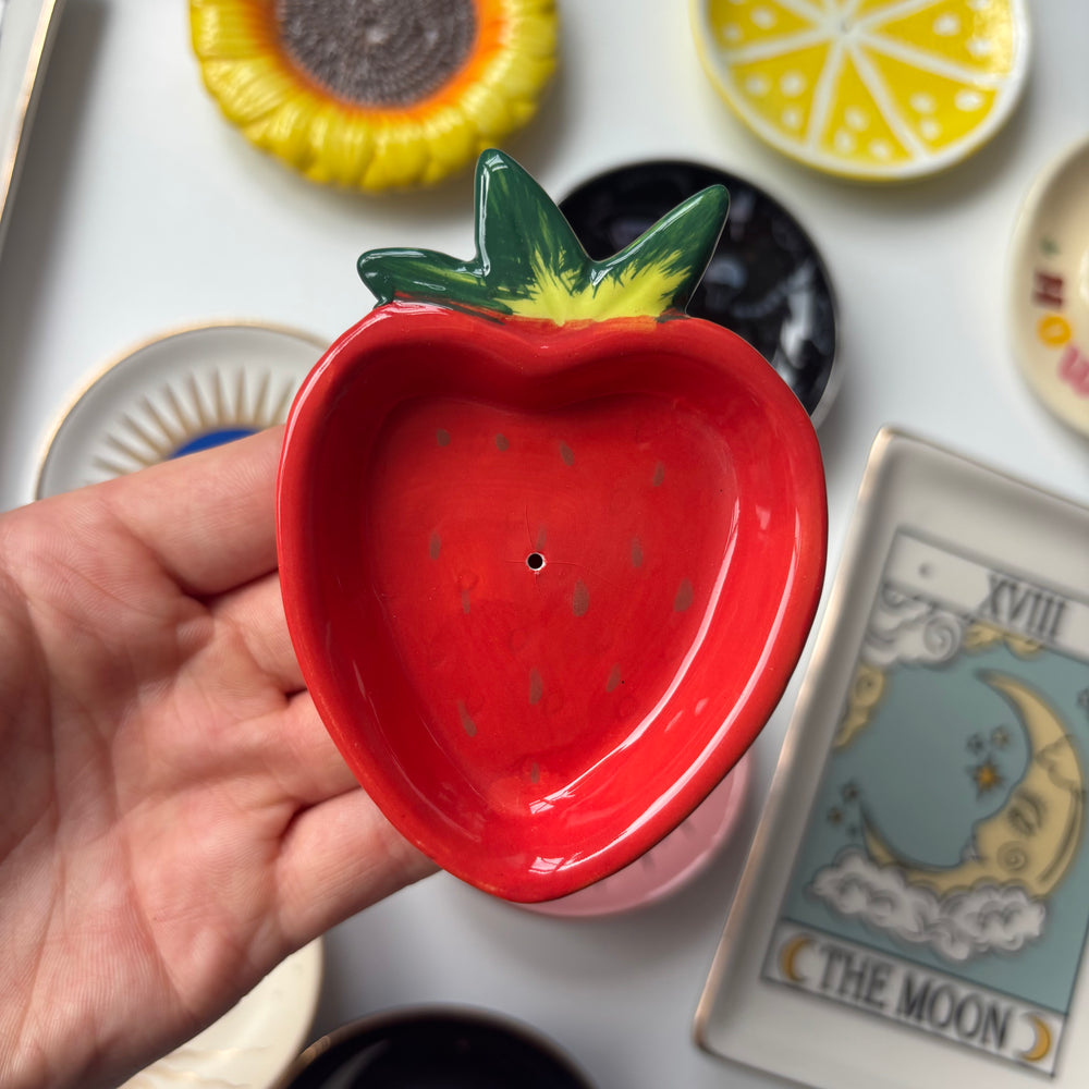 A strawberry-shaped Strawberry Incense Stick Holder being held in a hand. The glossy red dish has small green seed details, a green and yellow leafy top, and a small hole in the centre.