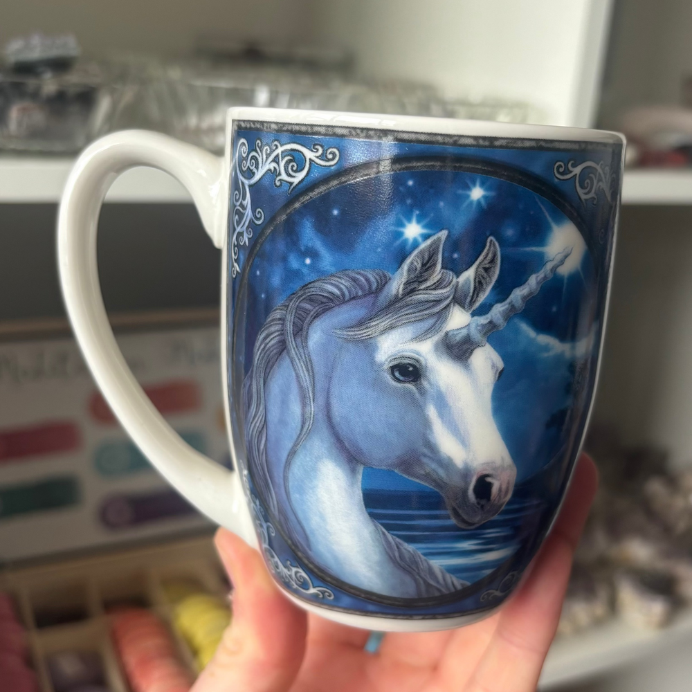 A hand holds a white ceramic mug featuring a detailed illustration of a white unicorn against a starry blue night sky background.