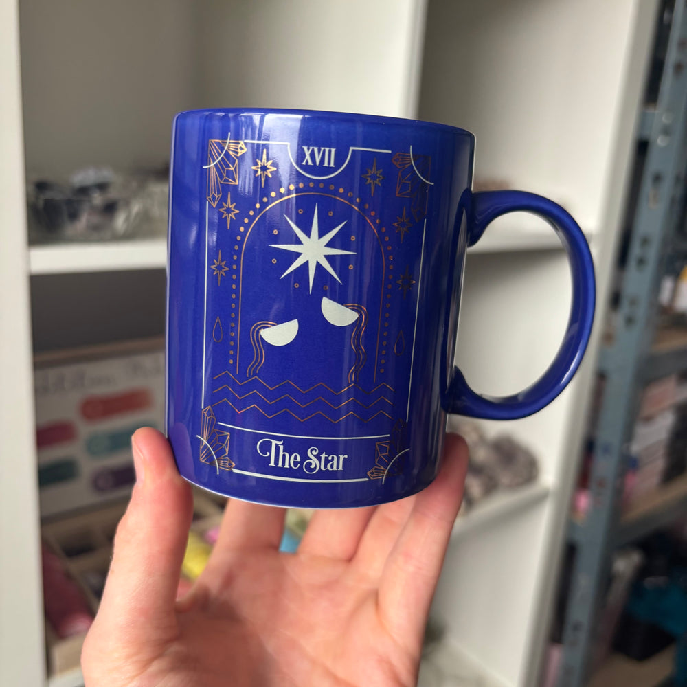 A hand holds a cobalt blue ceramic mug featuring a gold and white tarot card design labeled 'XVII The Star'.