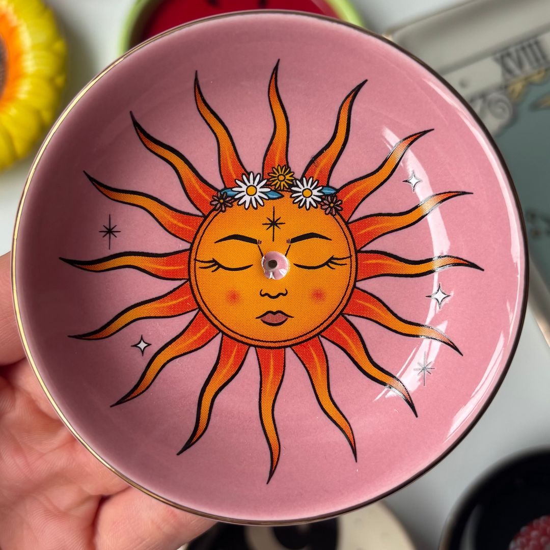 Hand holding a pink The Sun Celestial Incense Stick Holder against a background of various small dishes and tarot cards.