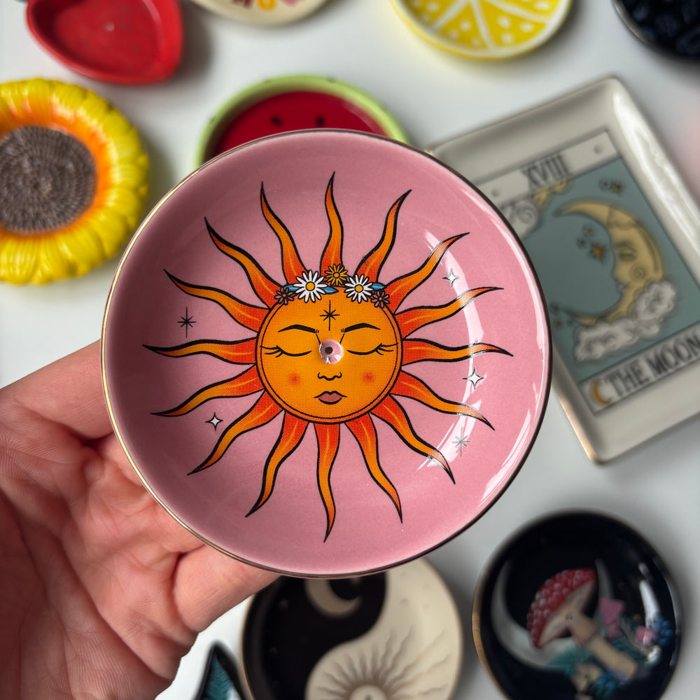 Hand holding a Sun Celestial Incense Stick Holder against a background of various small dishes and tarot cards.