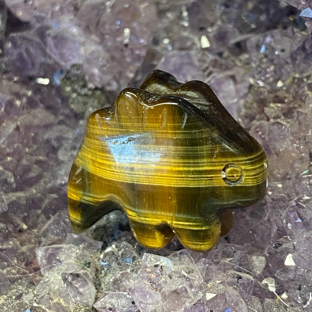 A carved tiger's eye Tiger Eye Stegosaurus Carving resting on a bed of purple crystal clusters.