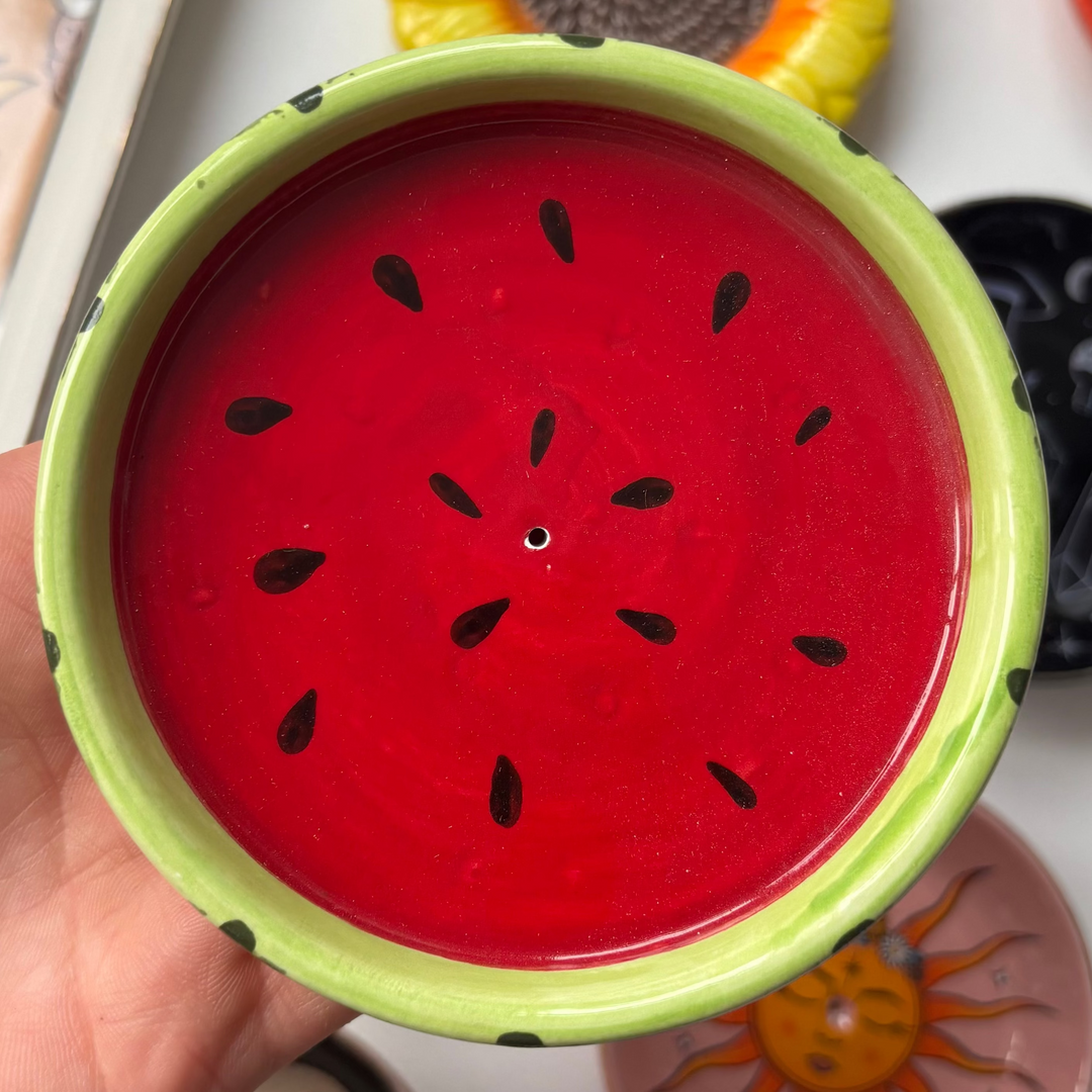 Hand holding a small red and green Watermelon Incense Stick Holder with a watermelon design against a background of various small ceramic items.