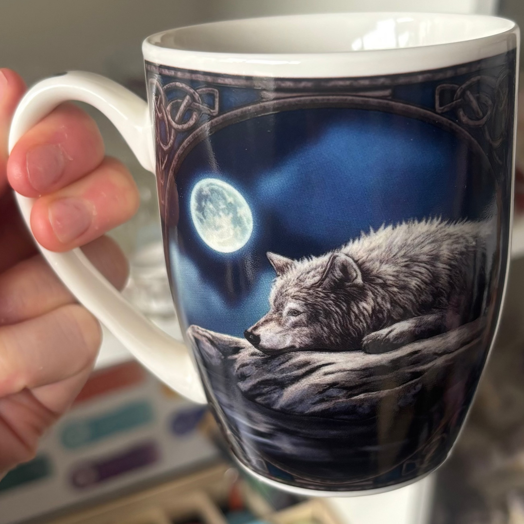 Mug with wolf design by Lisa Parker held by a hand, with a shelf and art supplies in the background