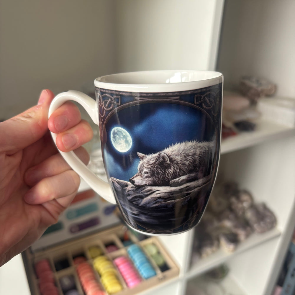 Mug with wolf design by Lisa Parker held by a hand, with a shelf and art supplies in the background