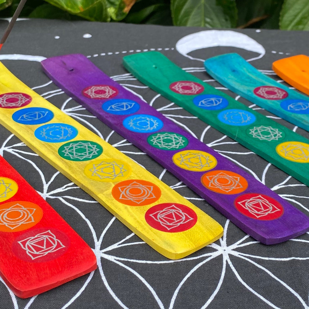 7 wooden incense in Red Yellow Purple Green Turquoise and Orange holders with seven colourful chakra symbols, shown with an incense stick on a black fabric background printed with a white flower of life pattern.