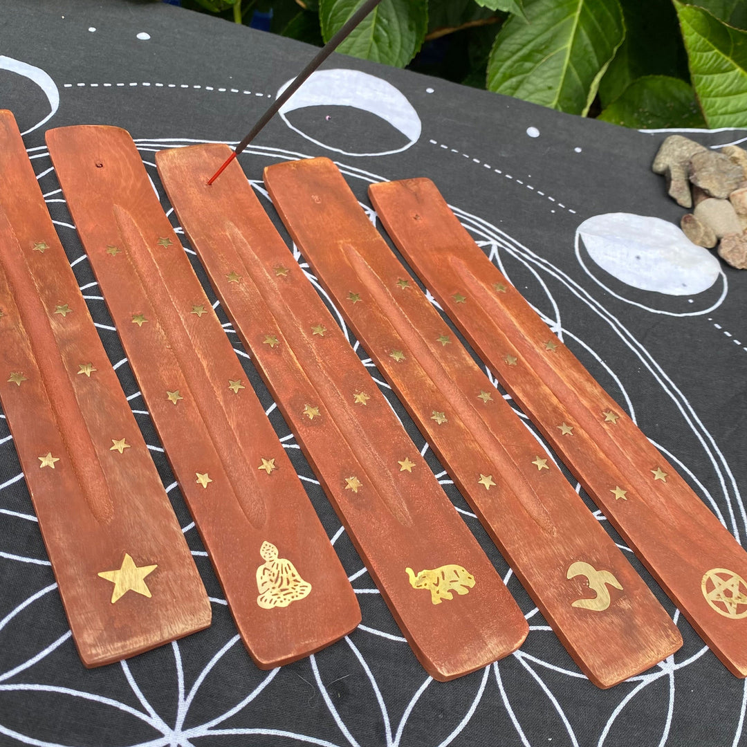 5 wooden incense holders with gold stars and Elephant, Om, Pentagram, Star and Buddha engravings and a burning incense stick, placed on a black fabric mat with a white geometric pattern, outdoors with green leaves in the background.