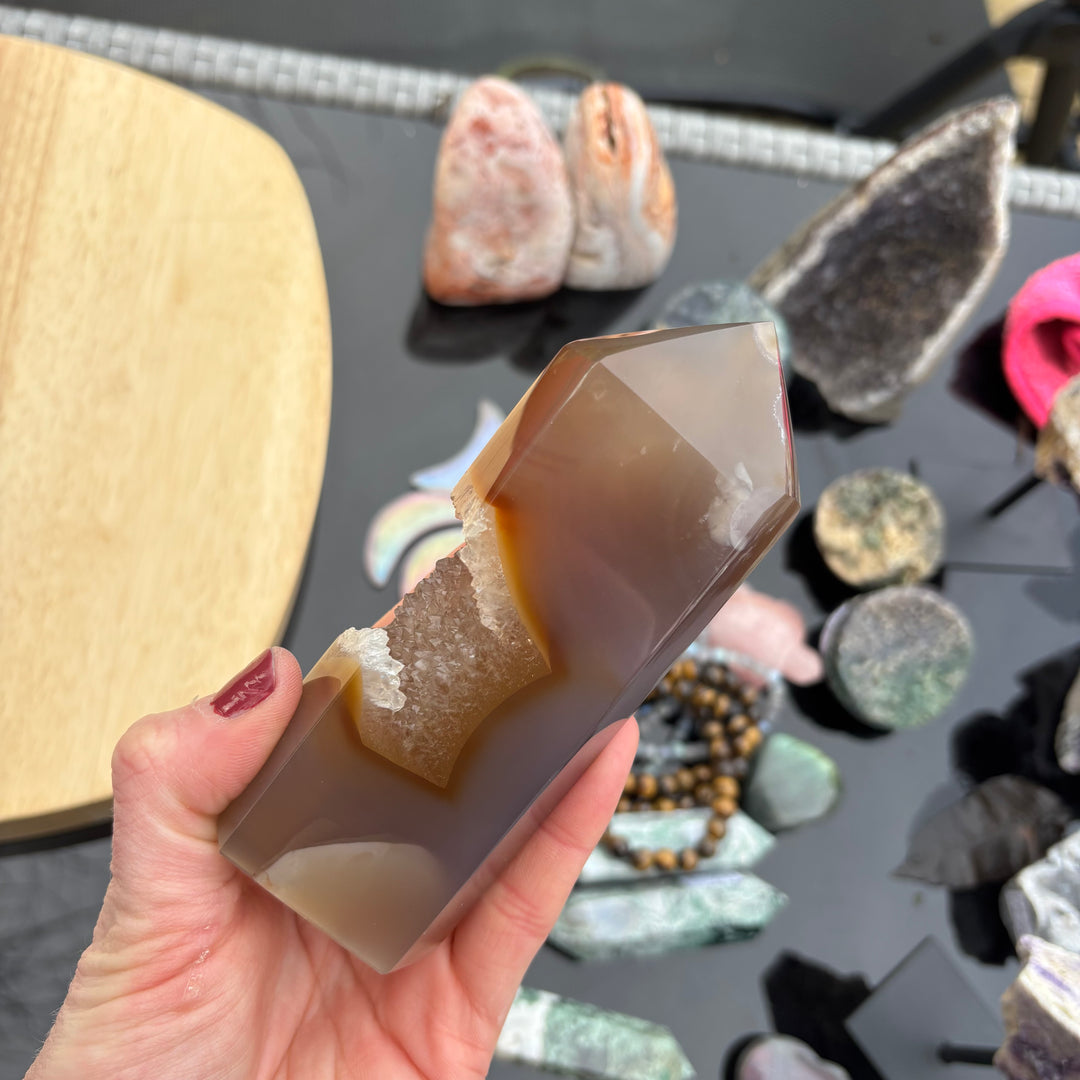 A polished Agate crystal tower held in an open hand, showcasing its smooth translucent brown faces, a sharp pointed apex, and a deep central cave lined with sparkling natural druzy crystals against an outdoor display backdrop.