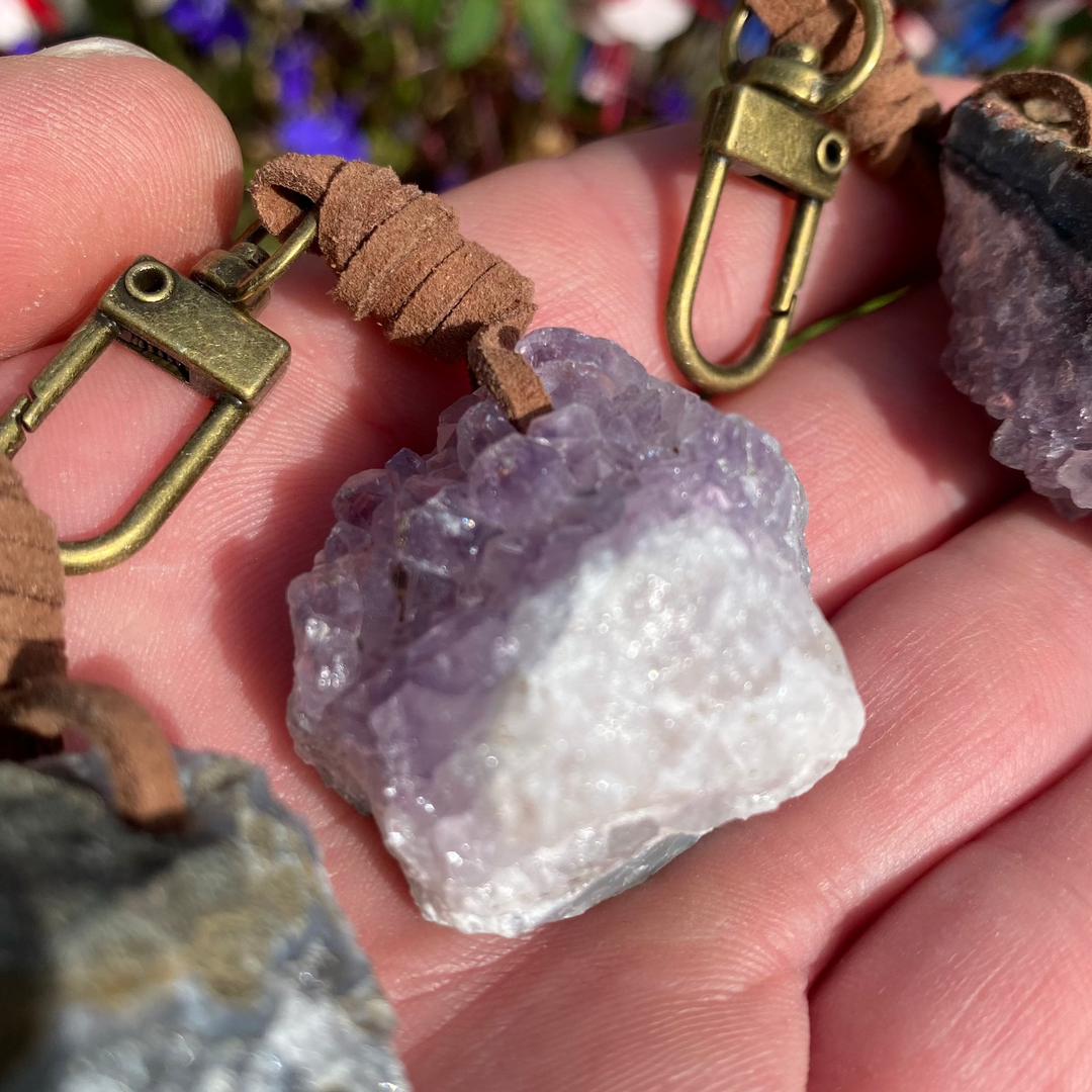 A detailed macro close-up view of a raw Amethyst crystal cluster keyring resting in an open palm. The shot highlights the rough, pale purple crystalline points, its natural rocky matrix backing, and a brown leather cord loop connected to an antique-style gold-toned brass lobster clasp.