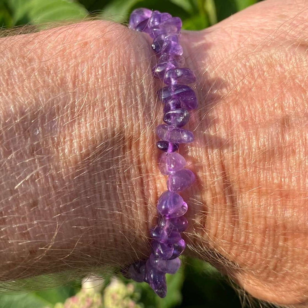 A direct vertical close-up shot of a person's wrist wearing the Amethyst crystal chip bracelet. The polished, irregular gemstone fragments glow with rich purple, violet, and lavender tones under bright, natural sunlight, contrasted against a soft-focus green garden background.