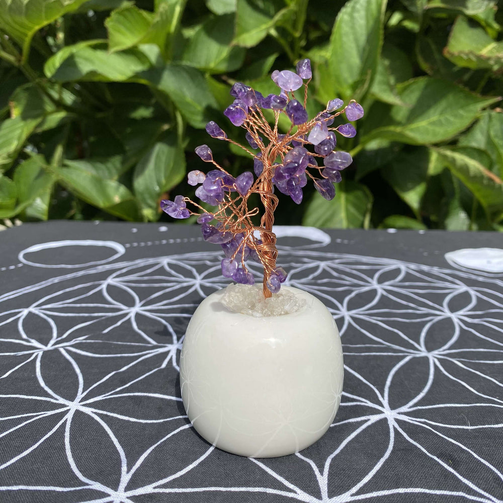 A straight-on outdoor shot displaying the complete profile of the amethyst gemstone tree on its white quartz base. The miniature crystal display is positioned against a vibrant backdrop of natural green foliage, emphasizing its elegant shape and calming decorative aesthetic.
