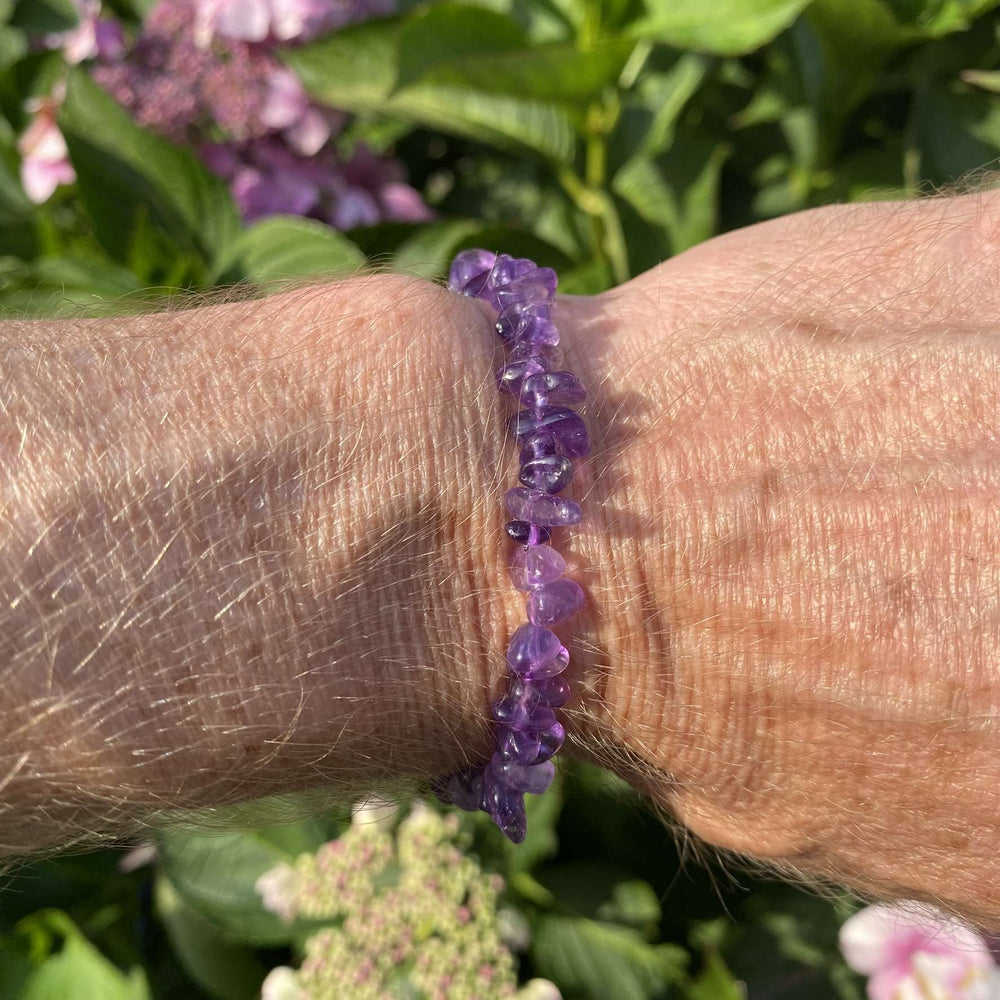 A side-angle outdoor view of the stretchy crystal bracelet worn on a wrist. The individual, deep purple amethyst chips catch the light beautifully, showing off their natural, glassy translucency against a textured background of green garden leaves and pale blossoms.