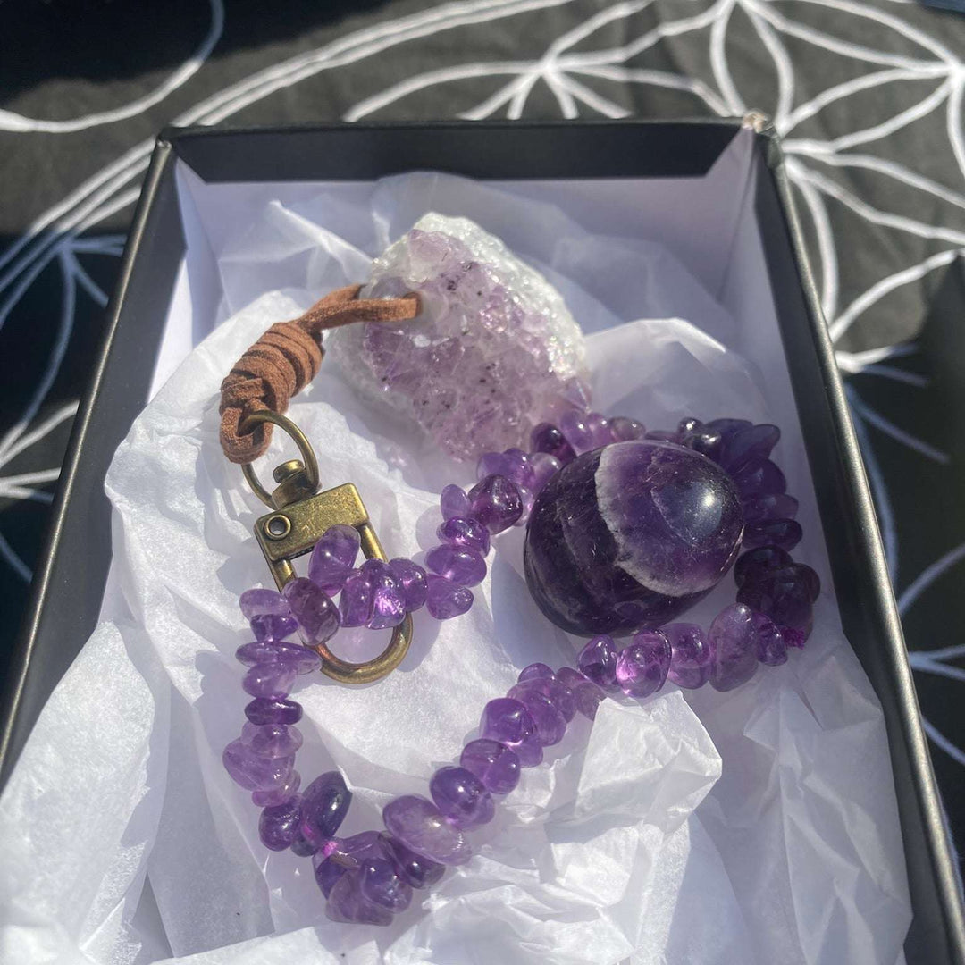 An open black cardboard gift box lined with protective white tissue paper, beautifully presenting an Amethyst crystal wellness set. Inside sits a polished purple Amethyst tumble stone, a stretchy gemstone chip bracelet, and a raw light purple Amethyst cluster keyring with a bronze metal clasp.
