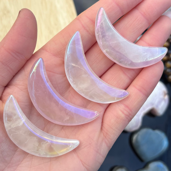 An open hand cradling four polished aura rose quartz half moon crystal carvings outdoors, showcasing their smooth crescent silhouettes, soft pink bodies, and shimmering metallic rainbow finishes for Heart Chakra alignment.