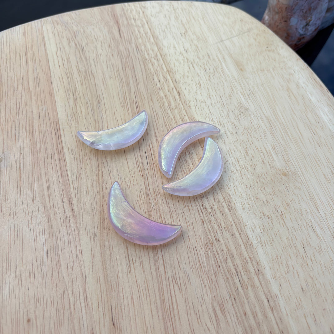 A flat lay shot of three aura rose quartz half moon crystal carvings arranged on a light wooden cutting board, displaying how the metallic sheen catches shifting daylight to add cosmic light to a vanity display.