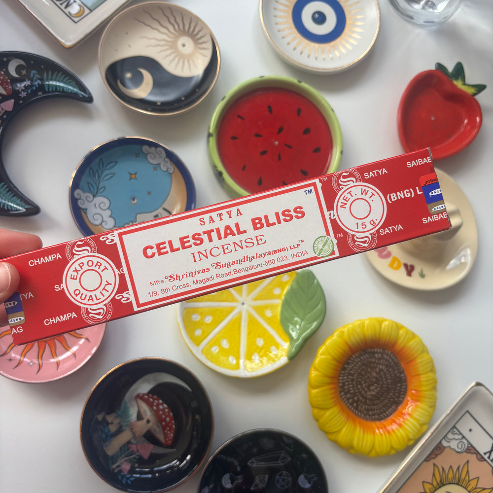 A high-angle overview layout showing a person's hand holding a 15g pack of Satya Celestial Bliss incense sticks on a white table surface. The colourful background collection of round ceramic ring dishes—featuring fruit designs, sunflowers, a crescent moon, and an evil eye emblem—creates a cheerful and whimsical retail layout under bright daylight.