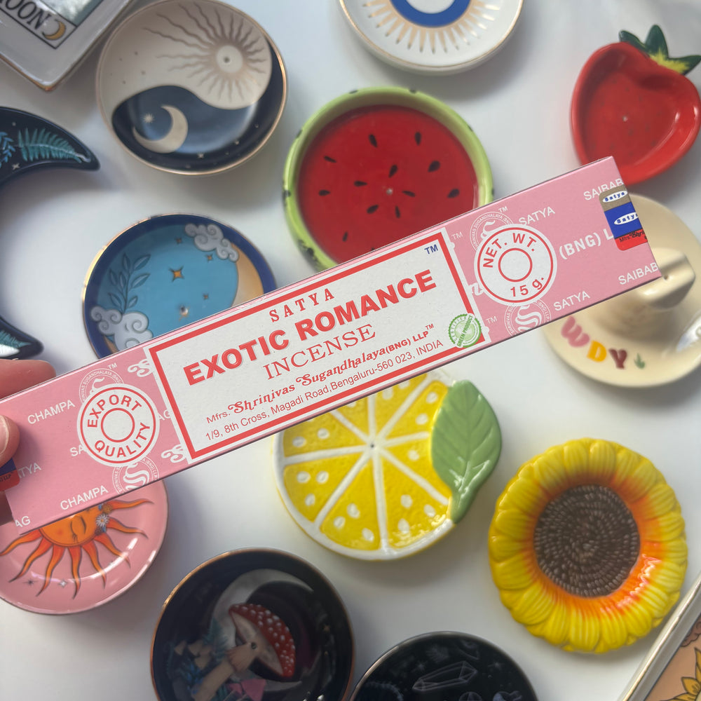 A high-angle overview layout showing a person's hand holding a 15g pack of Satya Exotic Romance incense sticks on a white table surface. The colourful background collection of round ceramic ring dishes—featuring fruit designs, sunflowers, a crescent moon, and an evil eye emblem—creates a cheerful and whimsical retail layout under bright daylight.