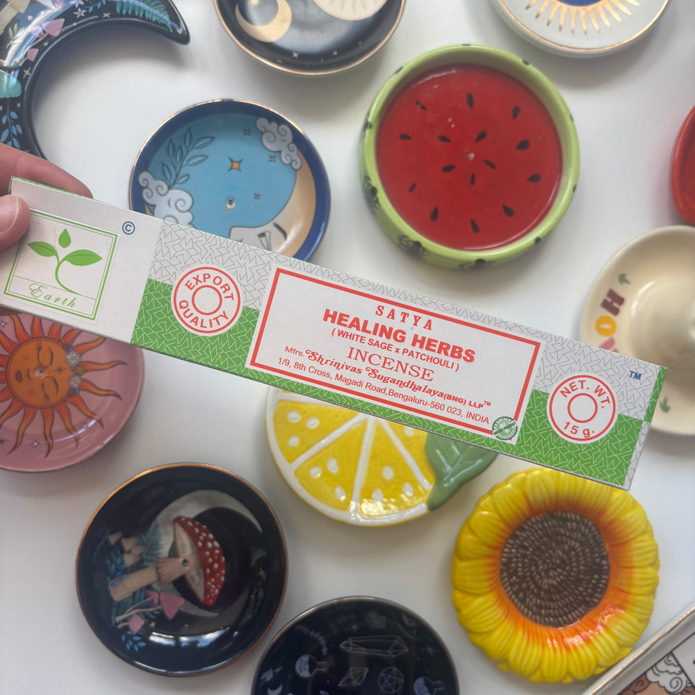 A high-angle overview layout showing a person's hand holding a 15g pack of Satya Healing Herbs incense sticks over a white table surface. The colourful background collection of round ceramic ring dishes—featuring fruit designs, sunflowers, a crescent moon, and an evil eye emblem—creates a cheerful and whimsical retail layout under bright daylight.
