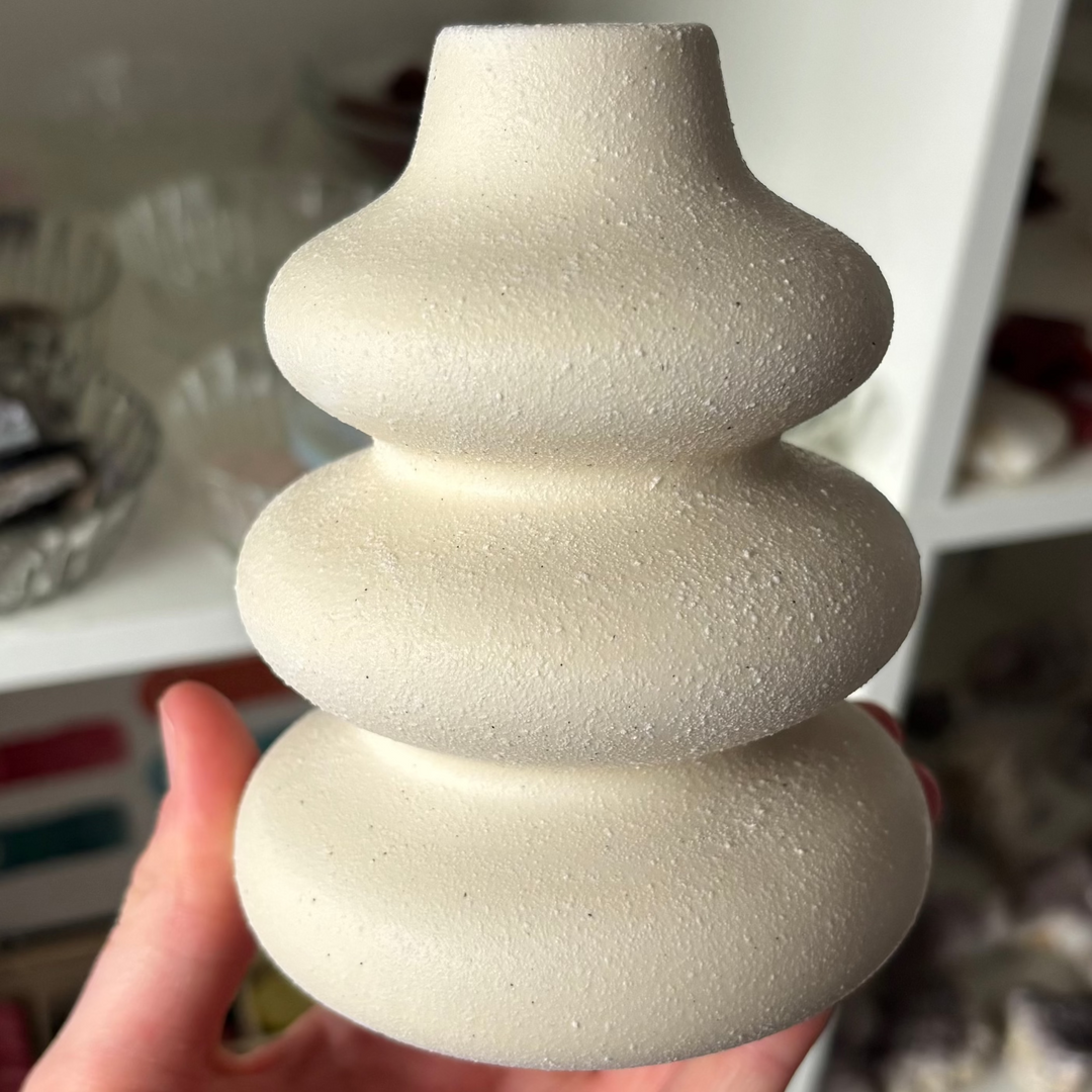 A direct close-up shot of a sculptural white ceramic single stem vase held gently in a hand. The minimalist design mimics three stacked, rounded river rocks with a narrowed, flared top opening, showcasing a finely textured sand-like matte finish against crystal shop shelving.
