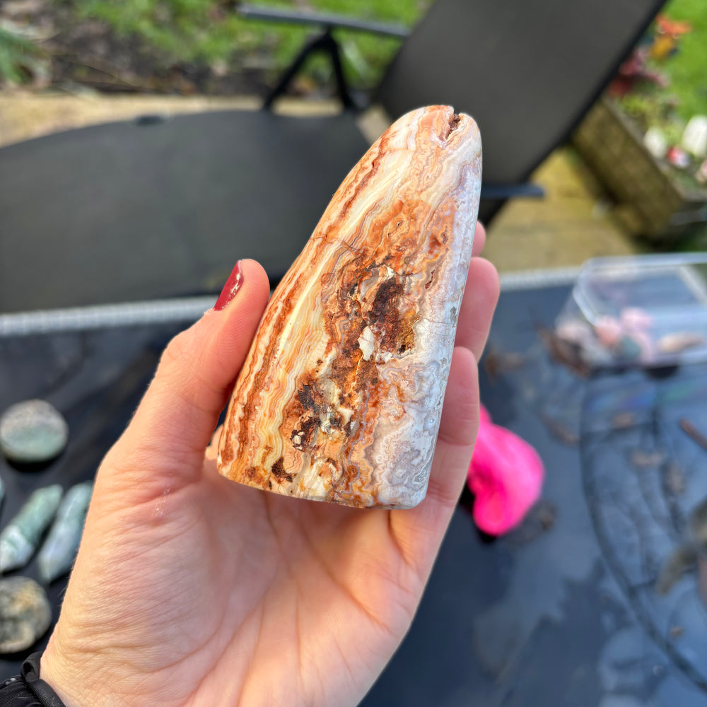 A hand holding the uniquely sculpted Cotton Candy Agate standing stone vertically, highlighting its sturdy flat base and bold, earthborn orange and white banding lines under daylight.