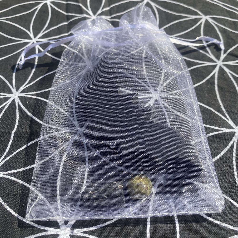 An overhead shot showing the complete gift set tucked inside a semi-transparent white organza bag, showcasing the silhouette of the black obsidian bat carving and the earthy tones of accompanying crystals, styled neatly on a sacred geometry patterned surface.