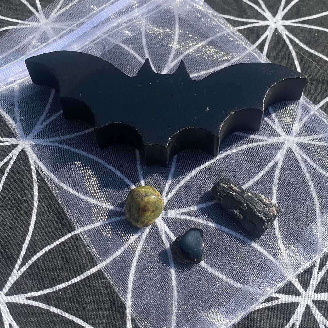 A close-up view looking down at a smooth, polished black obsidian bat carving arranged with three tumbled crystals on a sheer, white organza drawstring pouch, all styled against a dark grey background mat featuring a geometric white Flower of Life pattern.