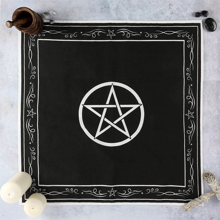 A square black cotton altar cloth laid out flat on a grey stone surface, featuring a white pentagram star inside a central circle and a flourished white border with small star illustrations, styled with white ritual candles and crystals.