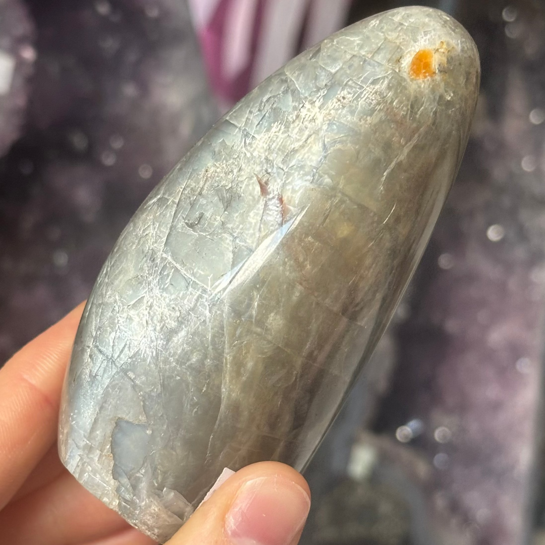 A close-up view of a polished Black Moonstone freeform crystal held gently in a hand. The stone showcases dark velvety layers, a soft silvery shimmer, and a small, natural peach-orange inclusions spot beneath its smooth, reflective surface.