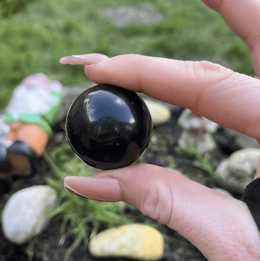 A close-up view of a highly polished Black Obsidian crystal sphere held carefully between a thumb and index finger outdoors, showcasing its smooth, glossy, midnight-black volcanic glass surface reflecting the light against a garden backdrop.