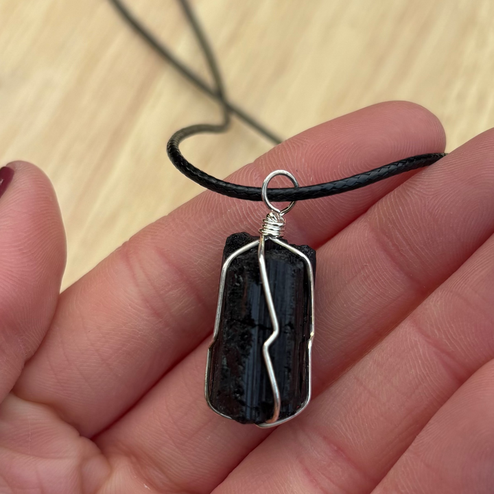 A hand holding a black tourmaline wire wrapped pendant necklace close up, showcasing the rugged natural texture of the raw protective stone cradled in delicate silver wire loops and suspended from a smooth black cord against a light wood background.