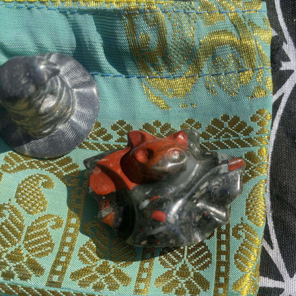 An angled close-up view of a polished crystal bat carving showing subtle green and red details next to the smooth rim of a Silver Obsidian witch's hat. The grounding duo is positioned near the edge of a golden patterned jacquard fabric pouch under bright, natural daylight.