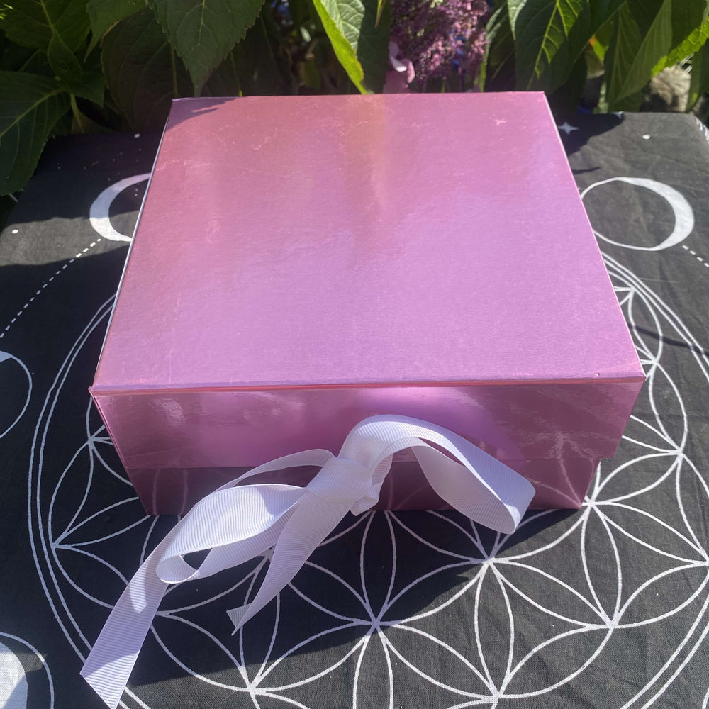 A close-up shot of the premium blush-pink cardboard gift box neatly closed and decorated with an elegant white satin ribbon bow. The packaging sits flat on a patterned black altar cloth outdoors under bright daylight, presenting an instantly gift-ready look.
