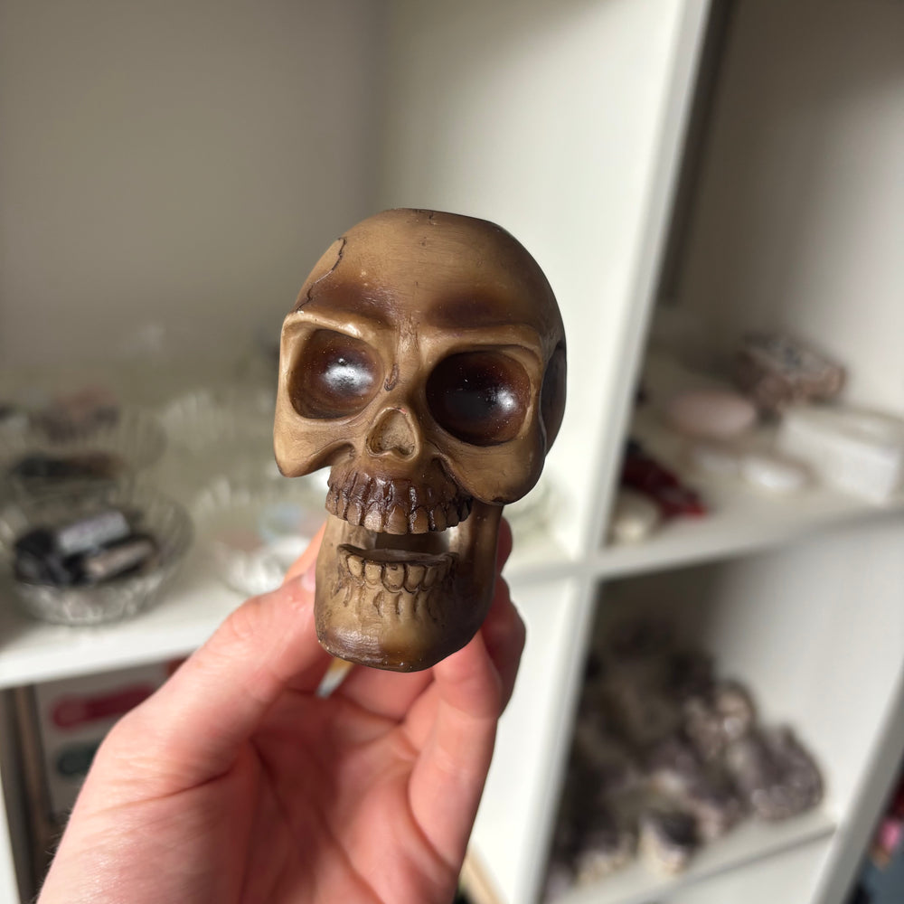 A high-angle view of a person's hand holding the bronze skull incense holder upright to showcase its compact 7.5cm height dimensions. The background displays softly blurred white boutique retail shelves, highlighting its structural layout for home altars.