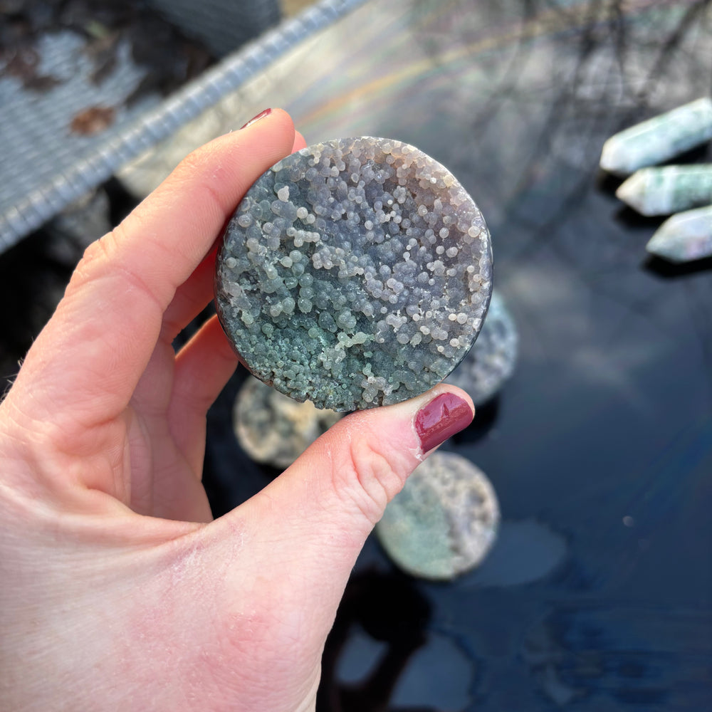 An outdoor shot showing the round purple and green Grape Agate gemstone chunk held in a hand from a distance, illustrating its compact, pocket-sized scale under natural daylight.