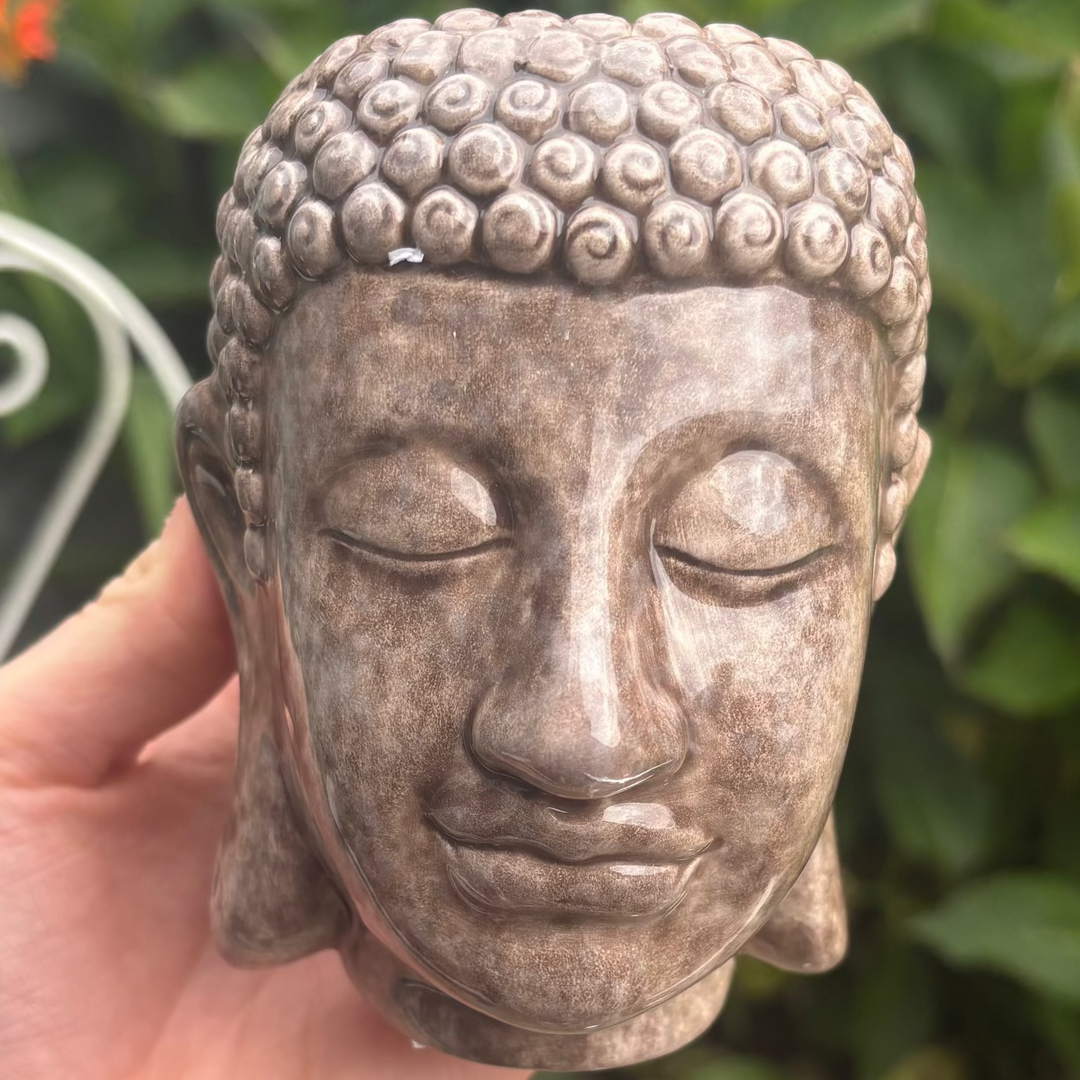 A direct macro close-up view of a ceramic Buddha head plant pot held gently in a hand. The pot features a beautifully distressed, textured brown stony finish with a serene facial expression, closed eyes, and tightly curled ushnisha hair details, captured outdoors against a soft-focus background of green leaves.