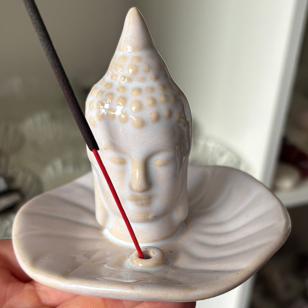 A close-up view of the white ceramic Buddha Incense Stick Holder held in a hand. The design features a smooth, texturised Buddha head standing in the centre of a curved ash catcher dish with a lit incense stick, perfect for anchoring a calm and protective energy.