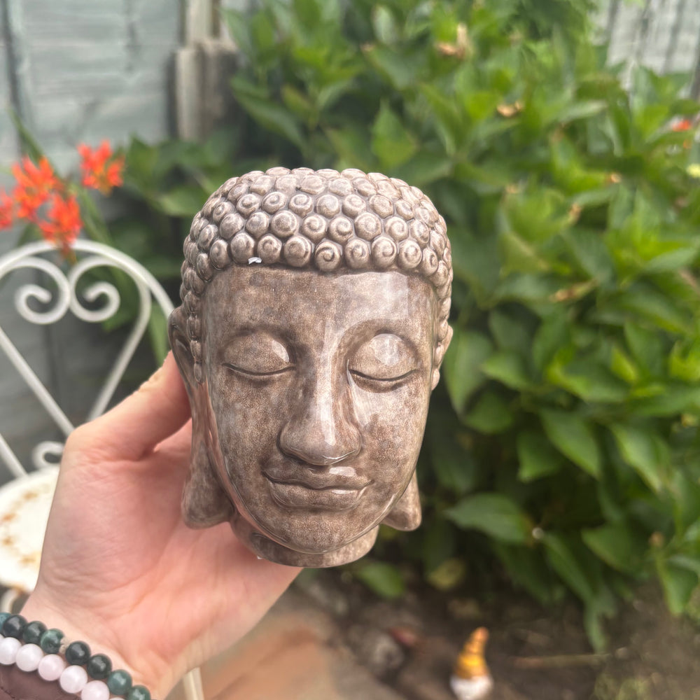 A high-angle outdoor shot of the ceramic Buddha head planter held up in a hand. The framing highlights its portable size and rustic finish, positioned next to a decorative white metal garden chair and vibrant green garden bushes under bright natural light.