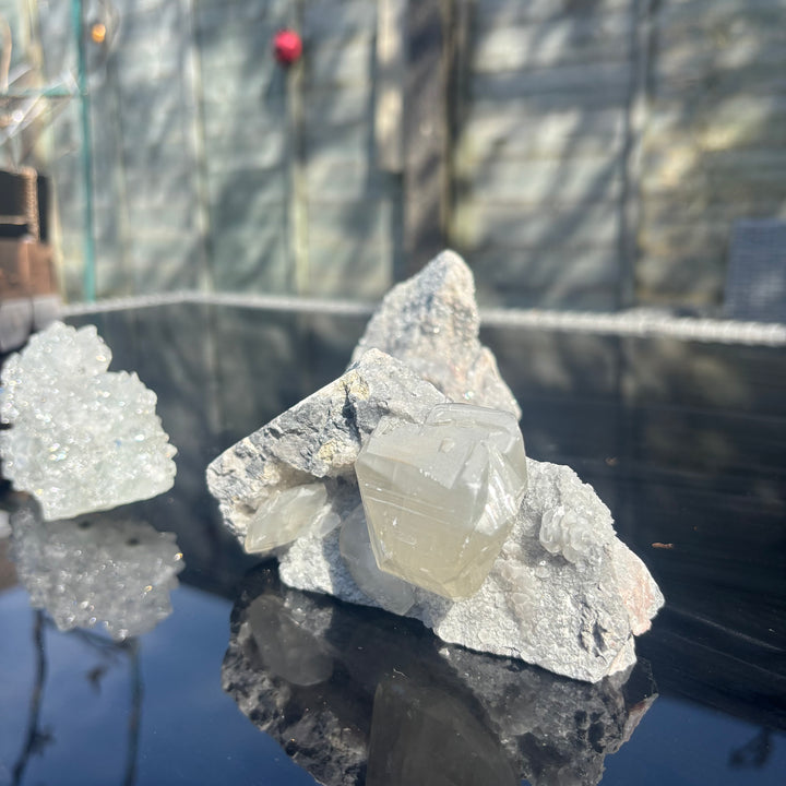 The rare dual-crystal specimen standing on a reflective outdoor table layout next to a smaller mineral fragment, highlighting how the frosty formations and contrasting textures filter energy and clear away mental clutter.