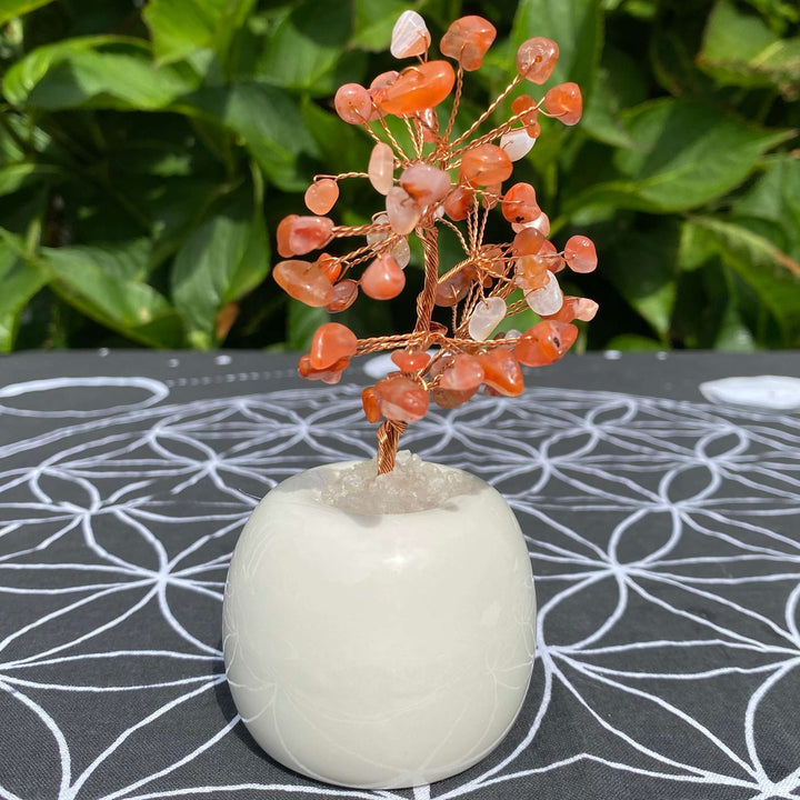 A handmade Carnelian crystal tree standing upright on a round, smooth white quartz base, displaying flexible copper wire branches laden with small, polished orange and red gemstone leaves on a black geometric mat outdoors.