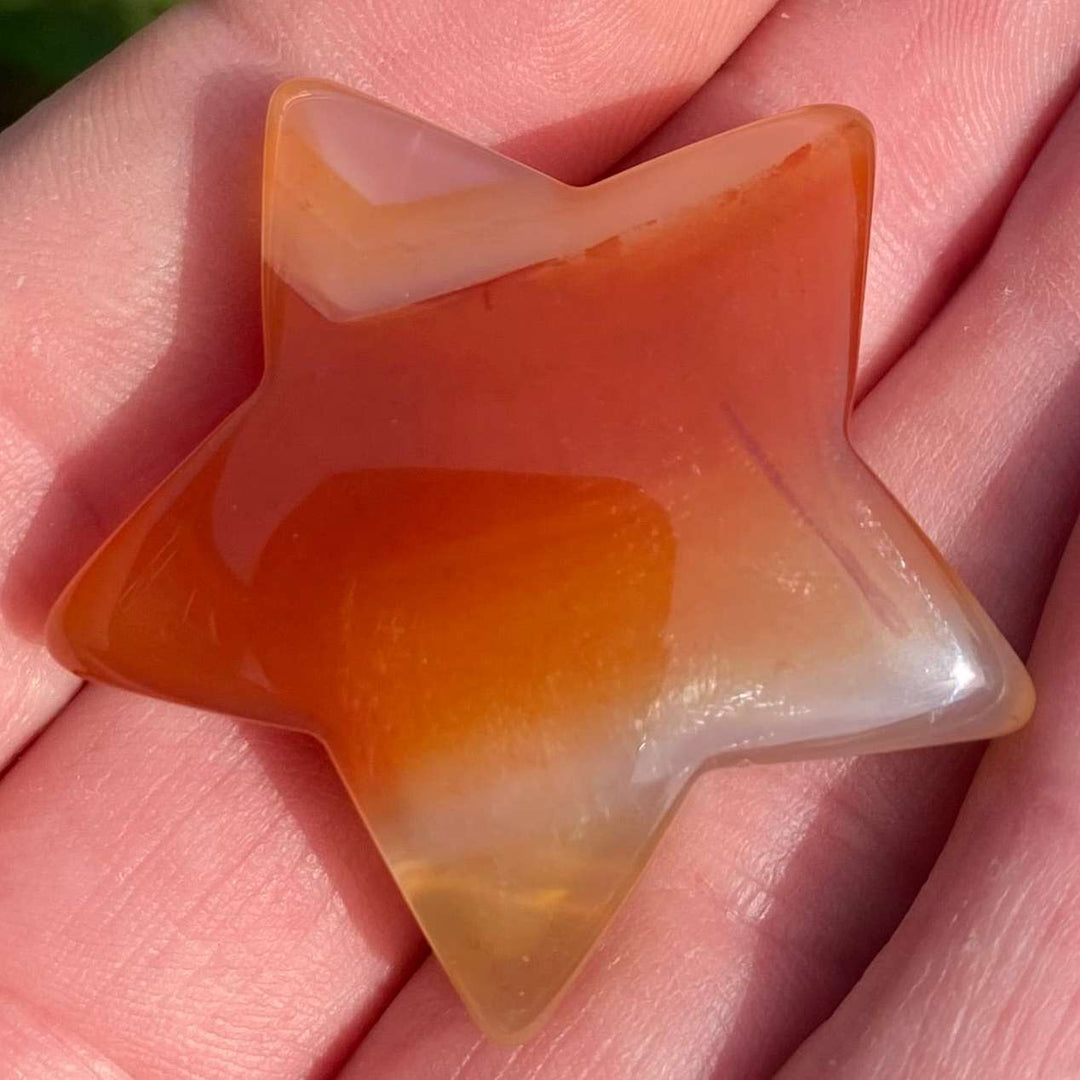 A macro close-up view of a polished carnelian drilled star crystal carving resting in an open hand. The celestial pocket gemstone showcases vibrant orange-red gradients, warm fiery swirls, and delicate translucent white banding across its glossy surface.