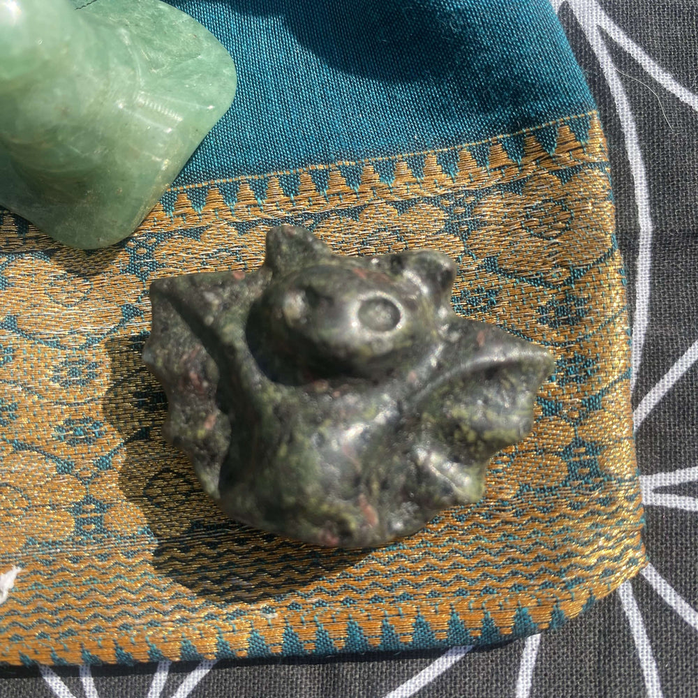 A close-up view highlighting the detailed wing carvings and dark, botanical patterns of the polished Moss Agate pocket bat figurine resting on a patterned fabric gift bag under daylight.