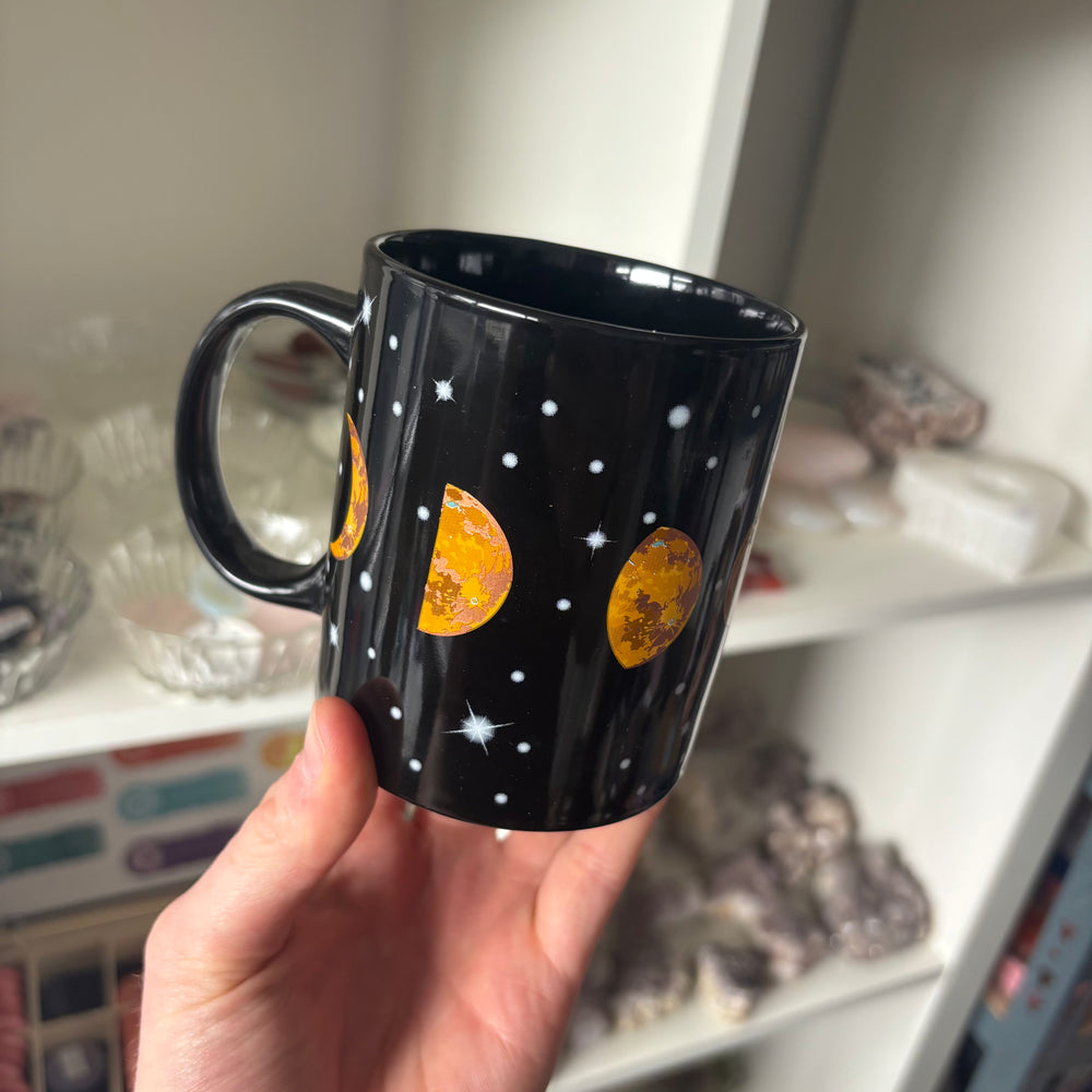 A high-angle view of a person's hand holding the black celestial mug to display its standard retail size and sturdy C-shaped handle profile. The background displays softly blurred white display shelves filled with crystals, showcasing the mug's eye-catching look for kitchen styling.